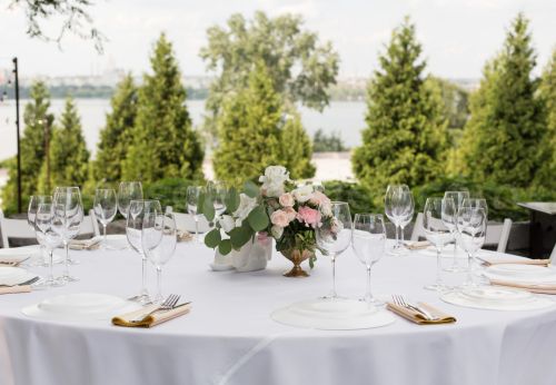 Table ronde dressée pour un mariage en extérieur, nappe blanche, verres à vin et centre de table en roses et eucalyptus, avec vue sur l'eau et les arbres