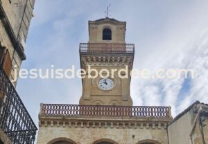 Tour de l'horloge de Vauvert Gard, en pierre, avec son cadran et ses balustrades sous un ciel nuageux